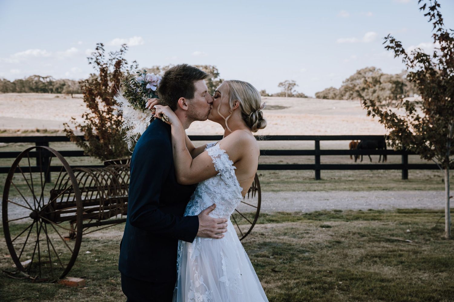 A romantic couple embraces and kisses in a rustic outdoor setting with wooden fences and natural landscape at sunset.