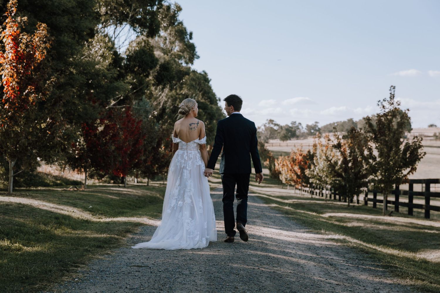 A couple in wedding attire walks down a tree-lined path during autumn with colourful fall foliage in the background.