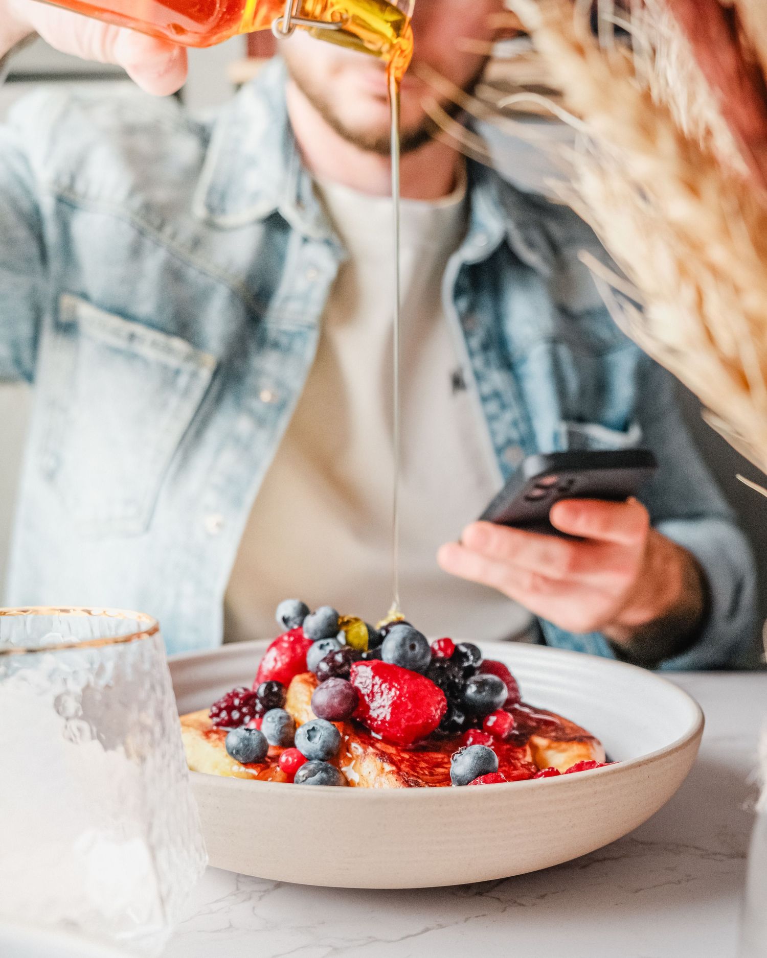 Plate of fruit and pancakes with a person pouring syrup on top with phone in their hand.