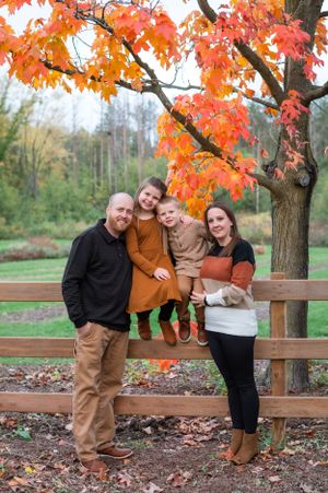 A happy family poses together by a wooden fence under vibrant autumn maple leaves.