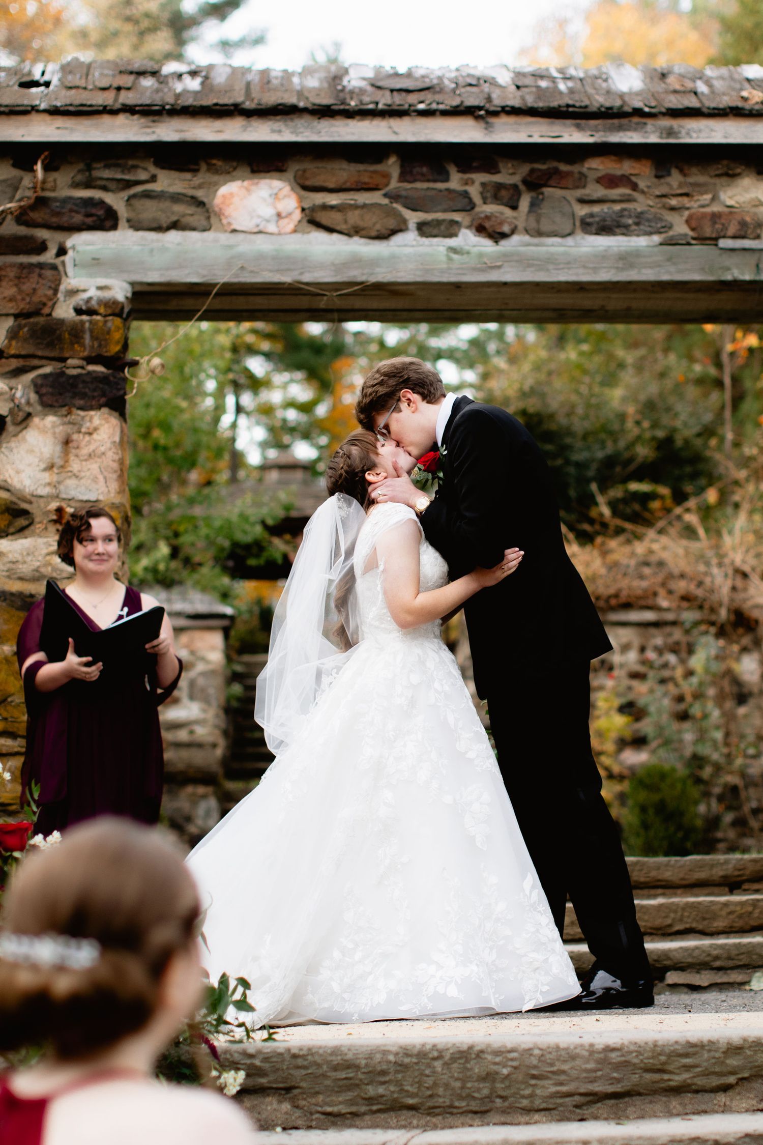 a wedding couple stands under a stone arch having their first married kiss; their officiant looks on, smiling