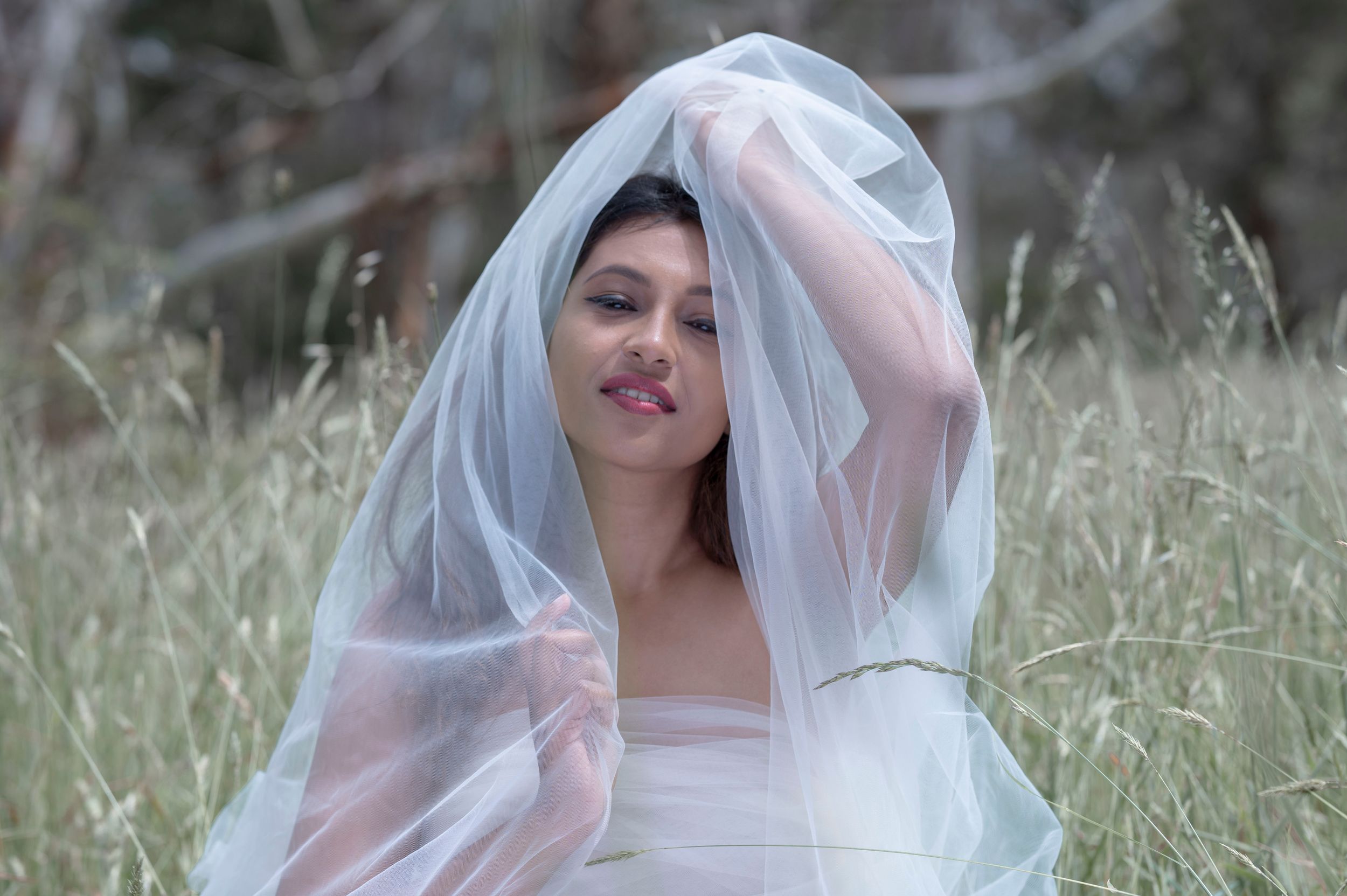Young woman in white dress lifting a sheer bridal veil while sitting in a grassy field.