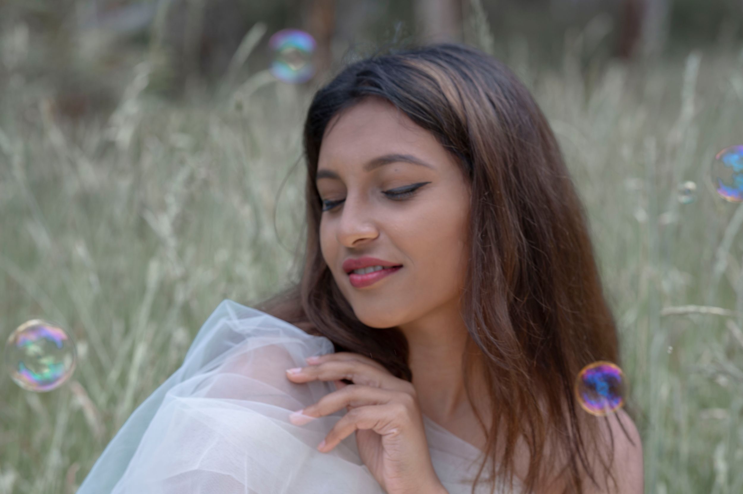 Beautiful young woman in white tulle dress watching colorful soap bubbles in a green field.