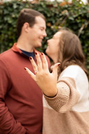 A close-up of an engagement ring being shown while a couple shares a joyful moment.