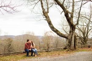A couple sitting close together on a wooden fence with bare trees and mountains in the background.
