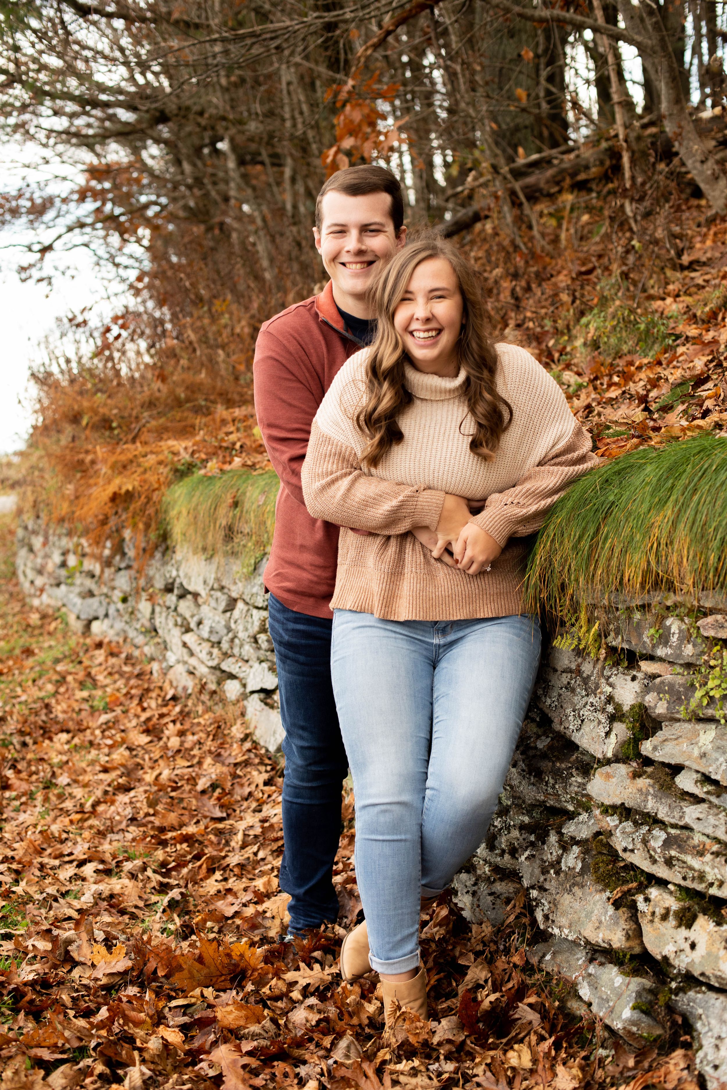 A couple embracing and laughing together against a stone wall surrounded by autumn leaves.
