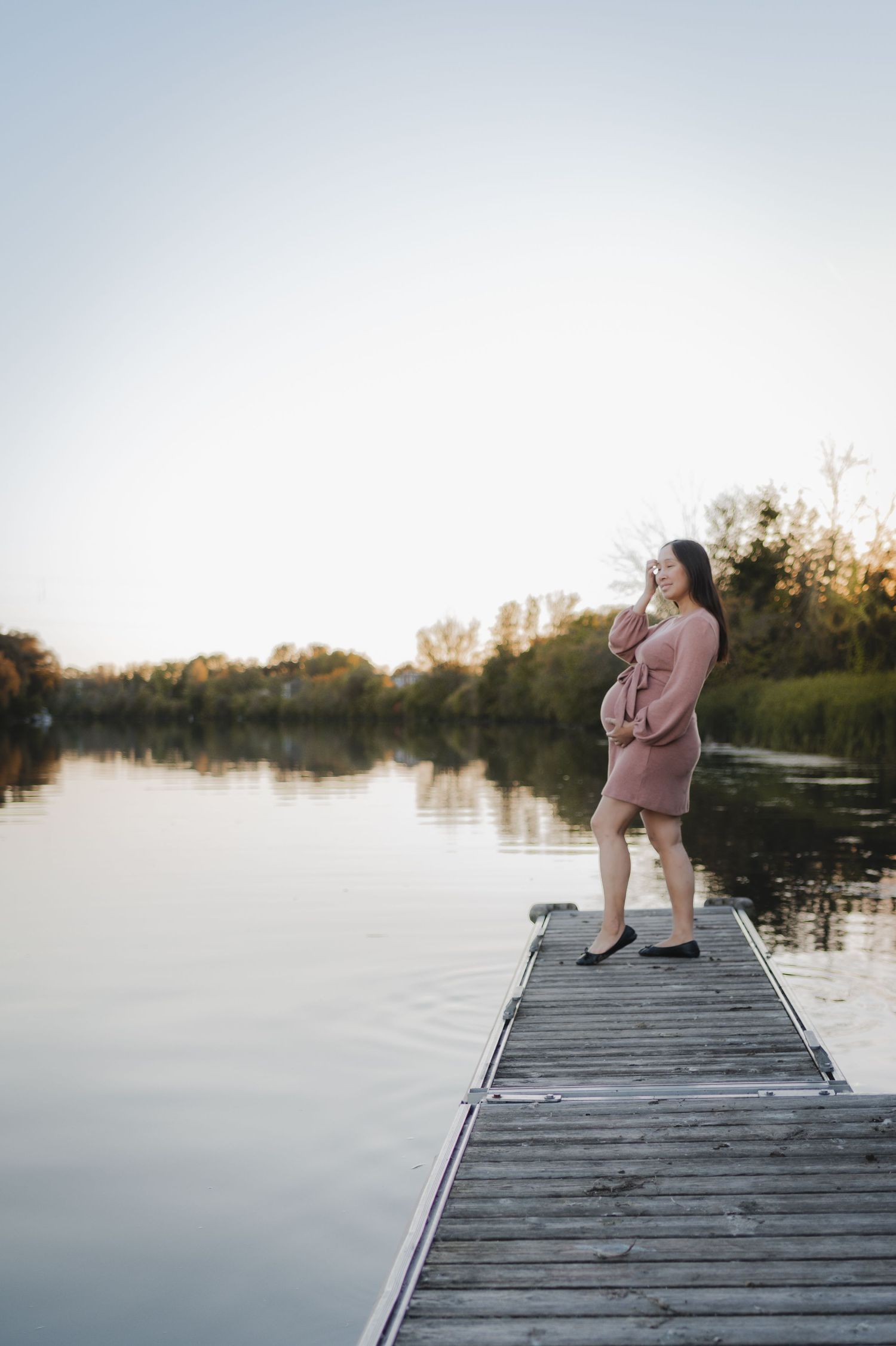 Person in pink outfit photographing sunset from wooden dock over tranquil lake.