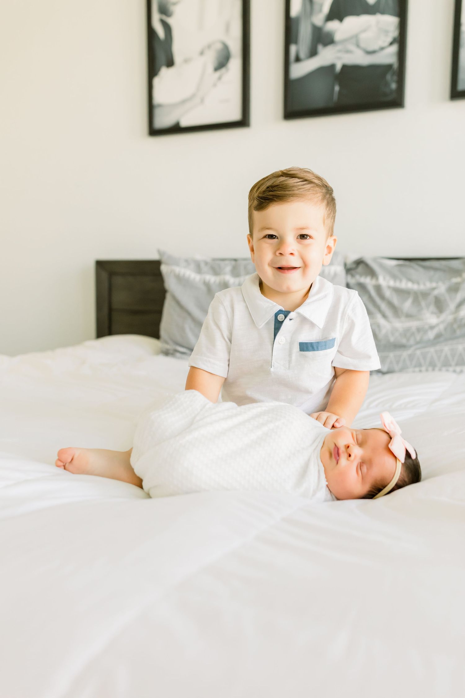 Two siblings share a sweet moment on white bedding with framed photos on the wall.