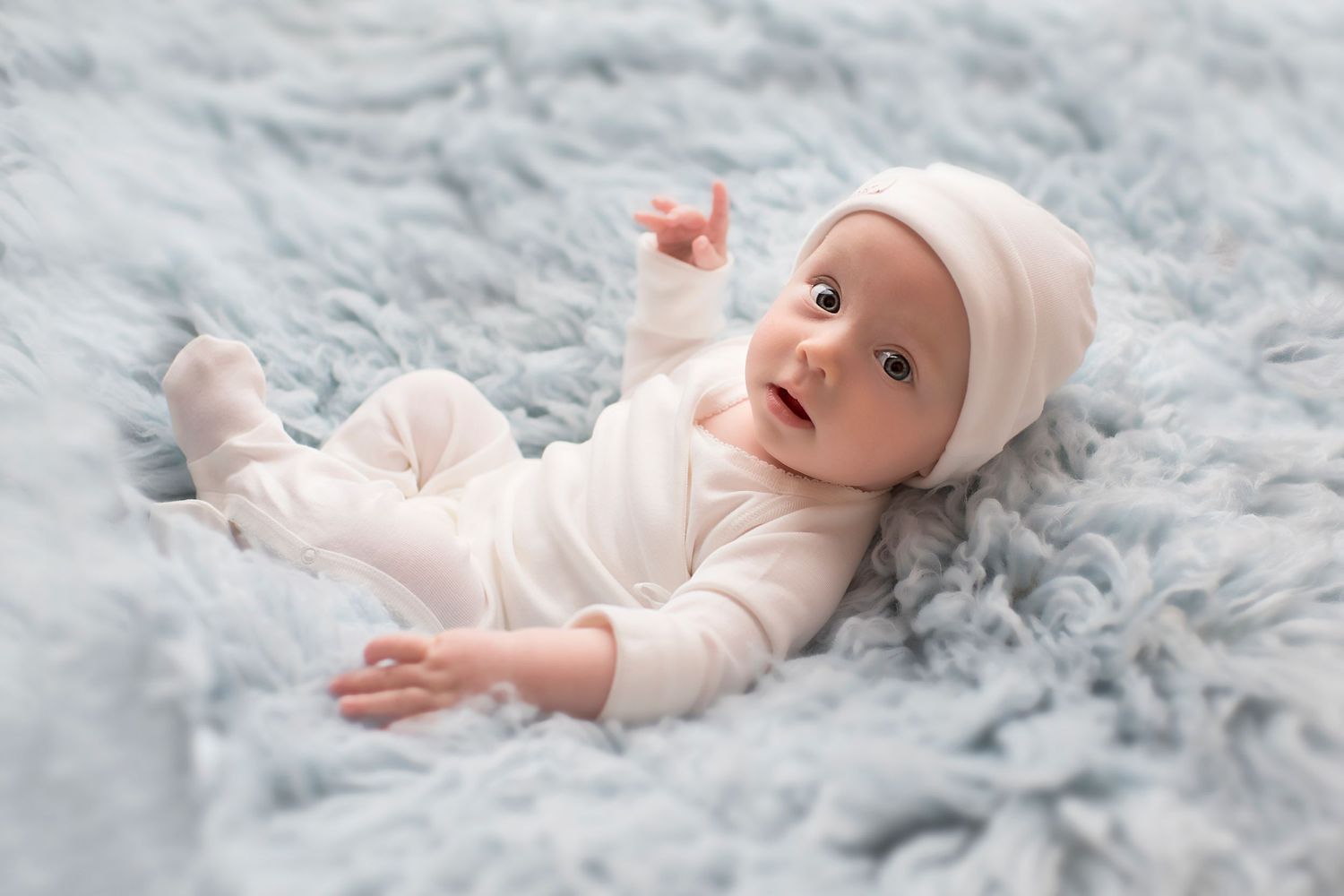Baby in white outfit lying on plush grey blanket during professional photoshoot.