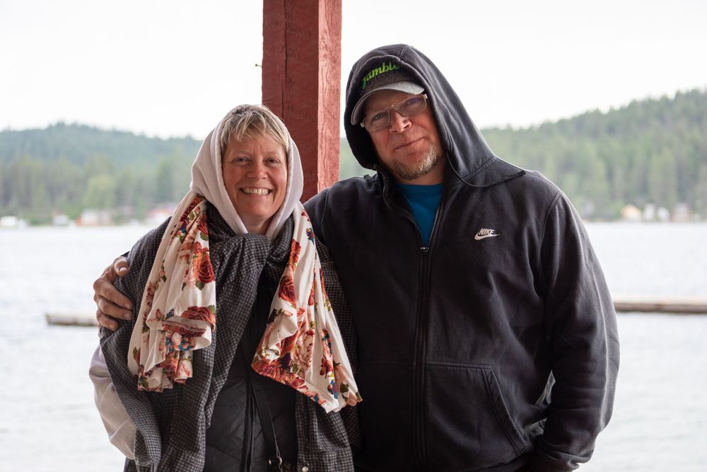 Two people wearing hooded jackets pose together near a lake.
