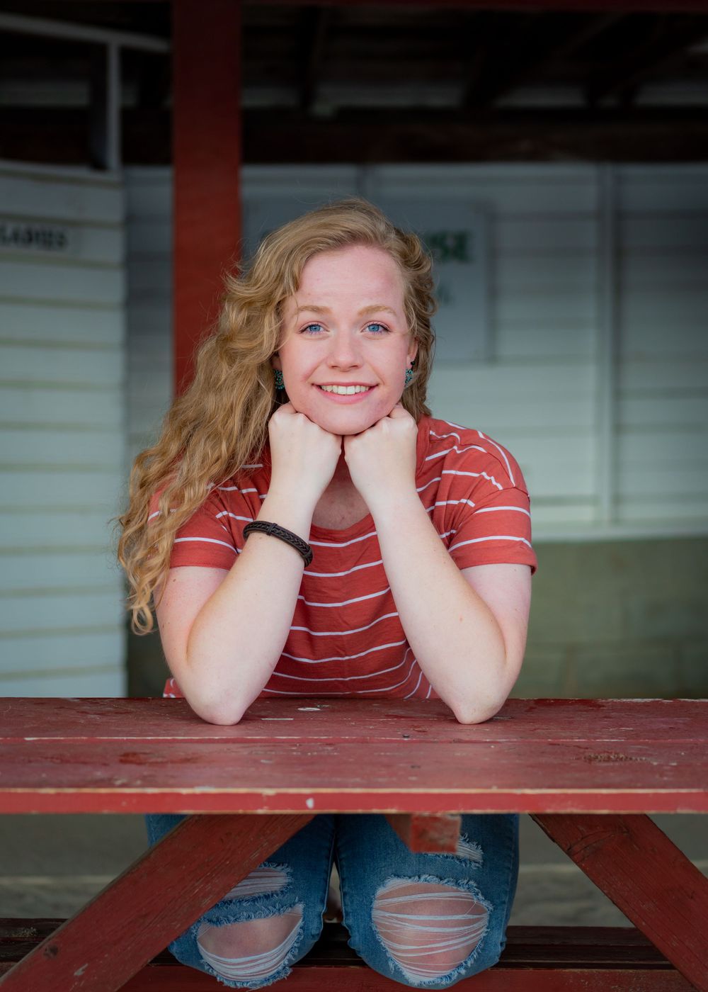 A casual portrait of someone in a red striped shirt and ripped jeans sitting at a red picnic table.