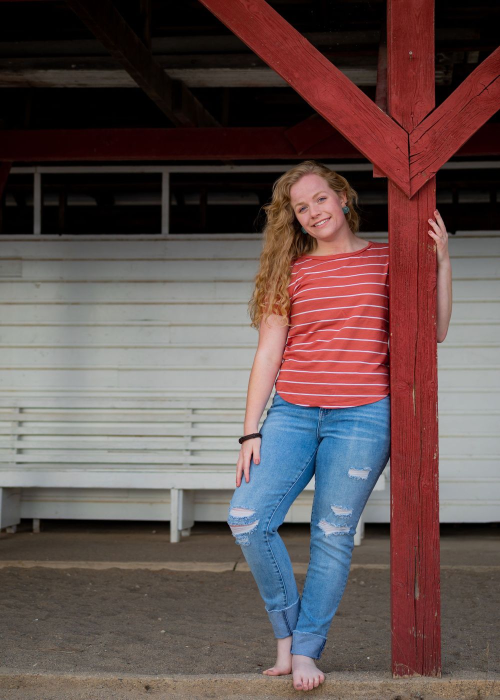 A casual pose against a red post wearing a striped shirt and distressed jeans.