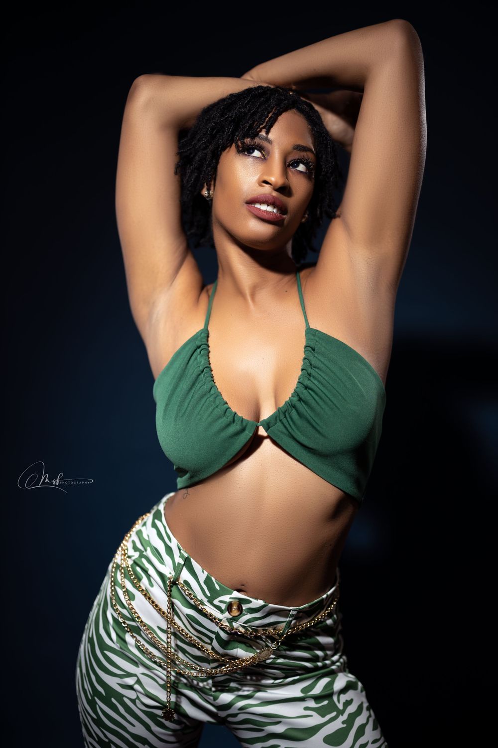 Fashion model in green crop top and animal print pants poses against dark background.
