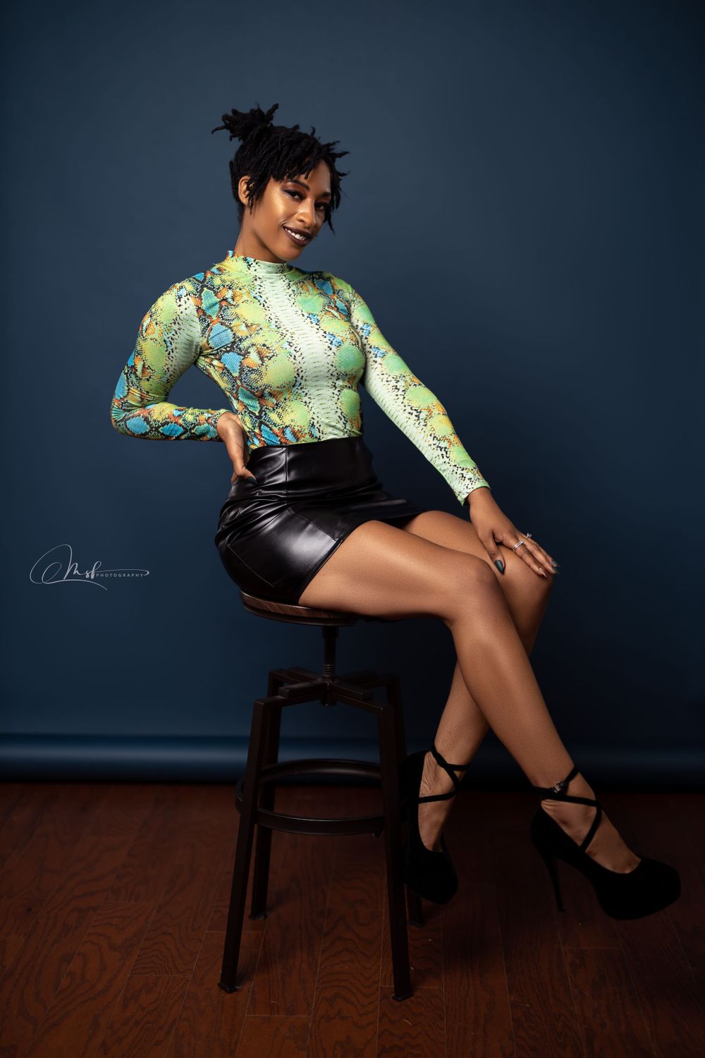 Fashion model in floral print top and leather shorts poses on wooden stool against dark blue backdrop.