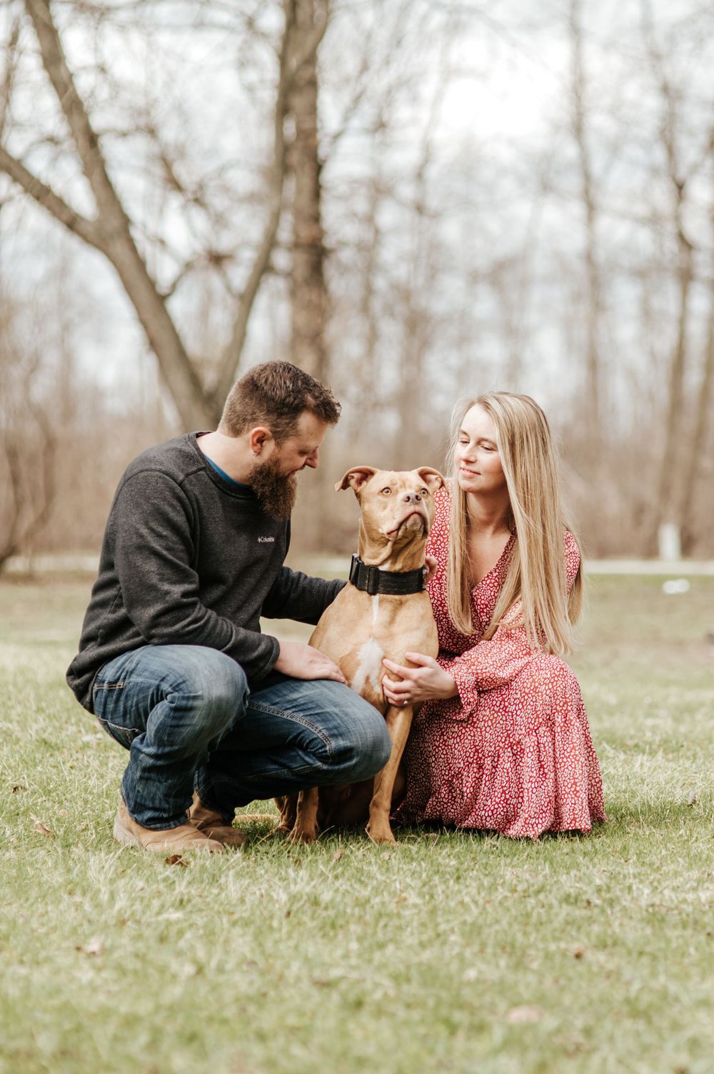 Couple sits on grass with their brown dog during outdoor photoshoot in wooded setting.