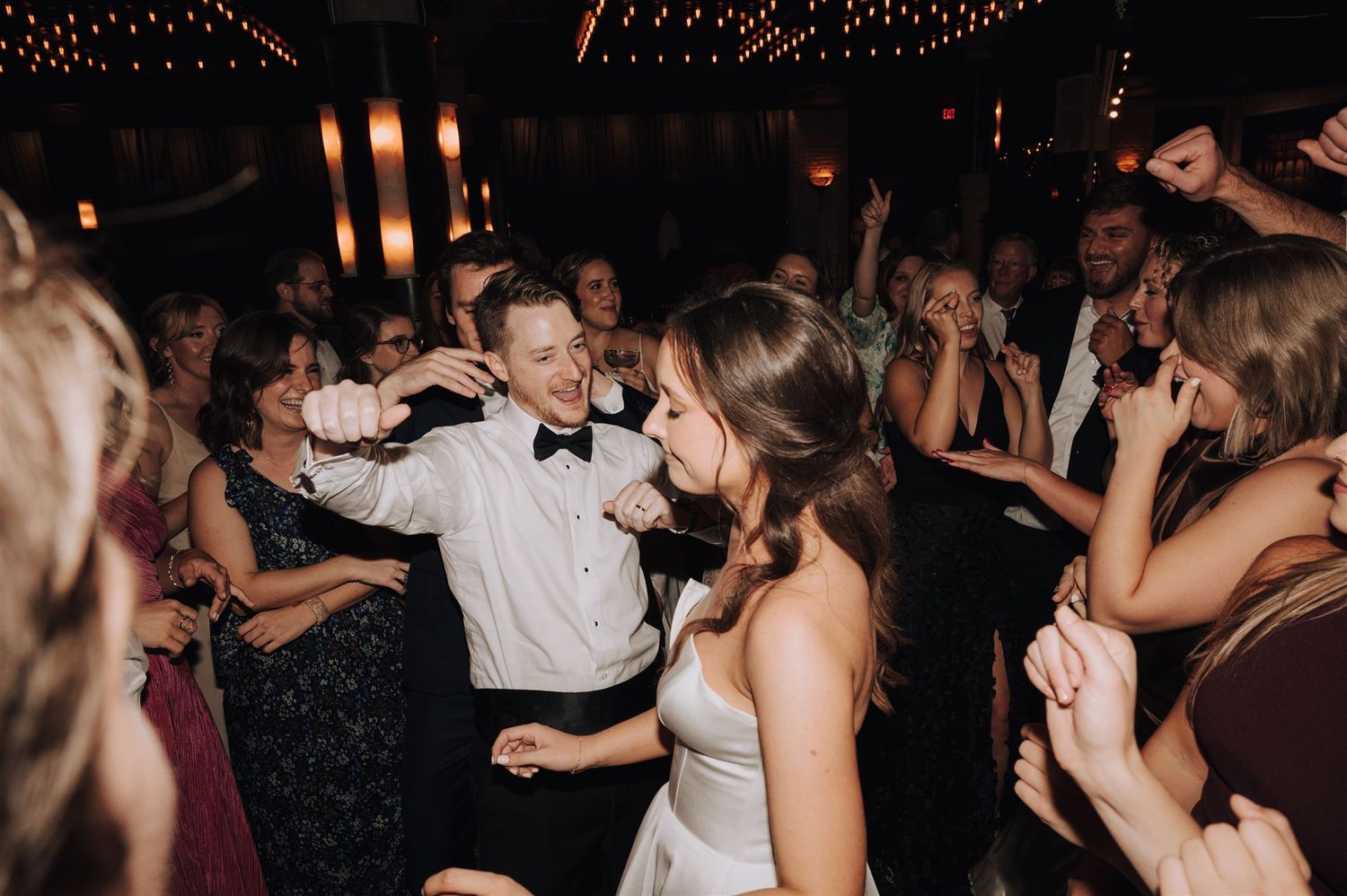 Wedding celebration on dance floor with guests dancing under warm string lighting.