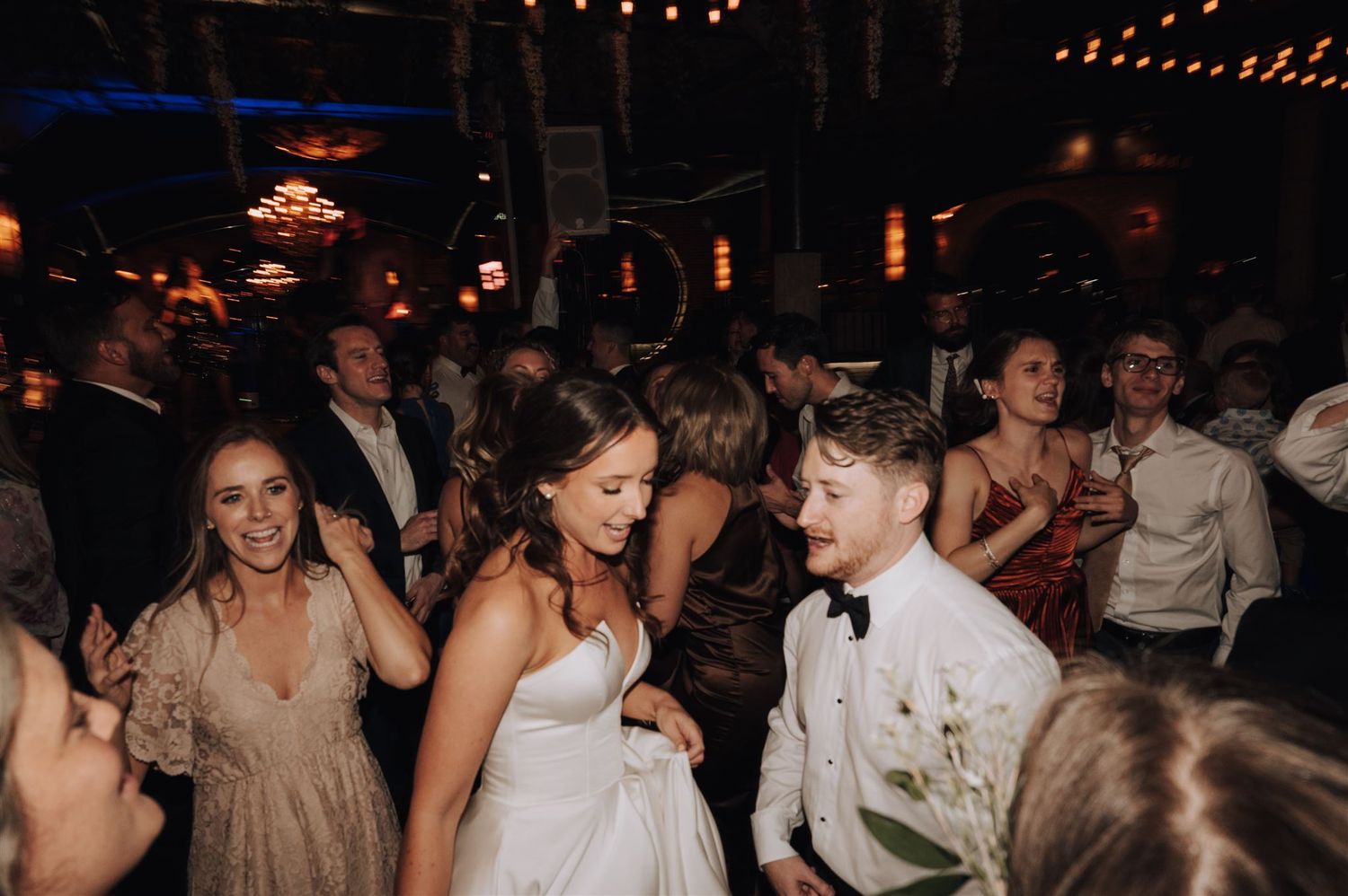 Wedding guests dance and celebrate on a dimly lit dance floor with string lights overhead.