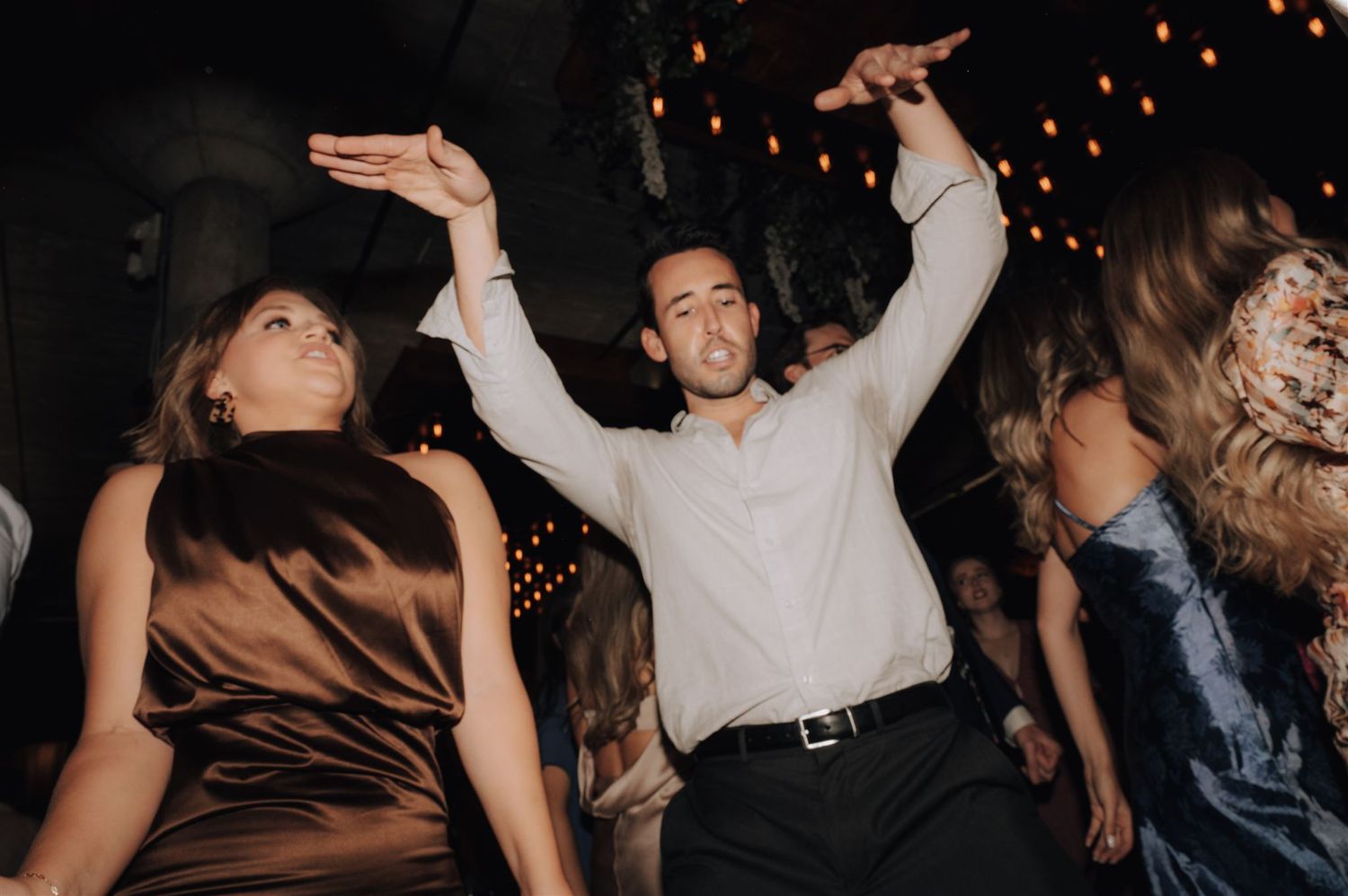 Energetic dancers with raised arms moving to music under ambient string lights.