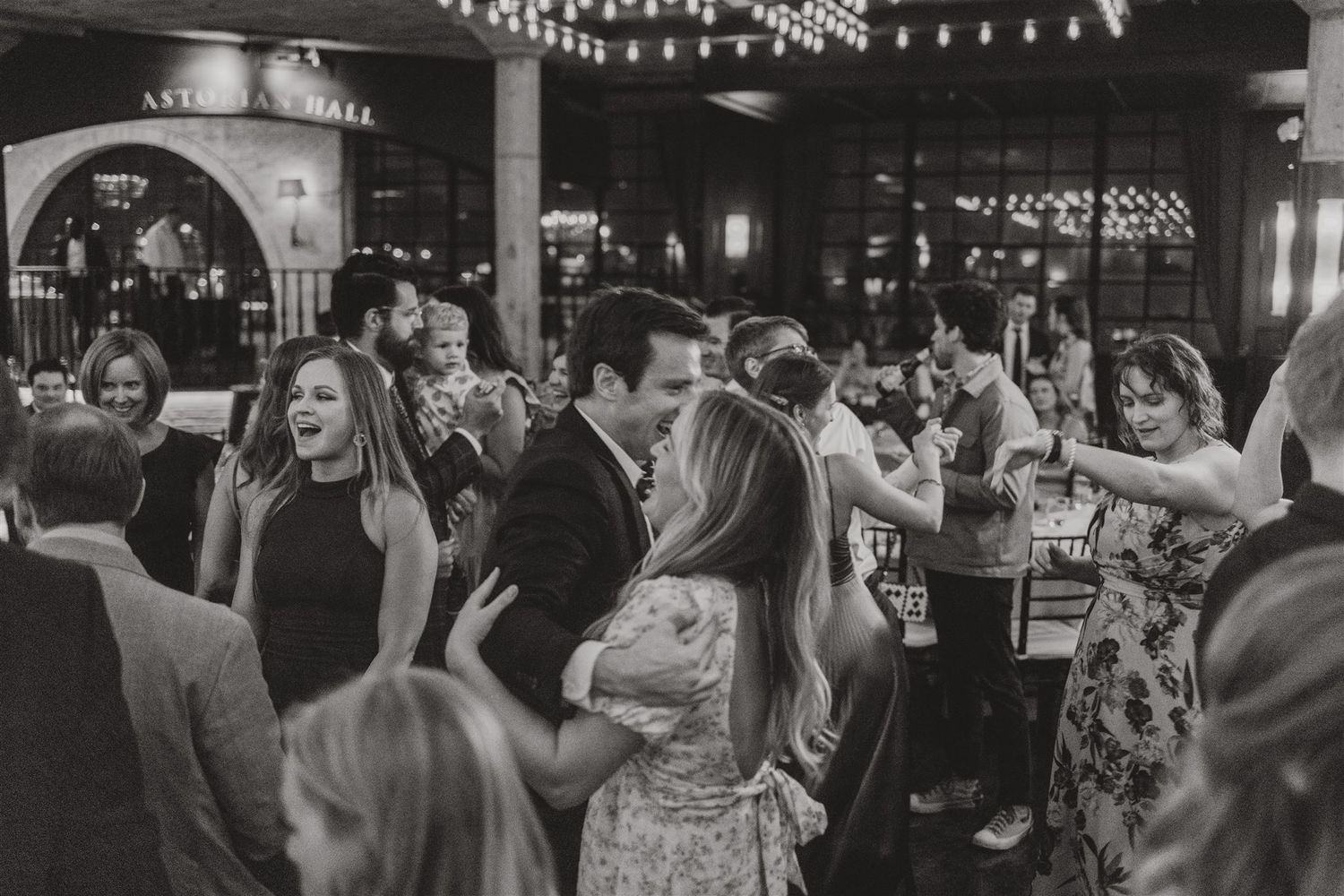 Black and white photo of a crowded wedding reception with guests mingling under string lights.