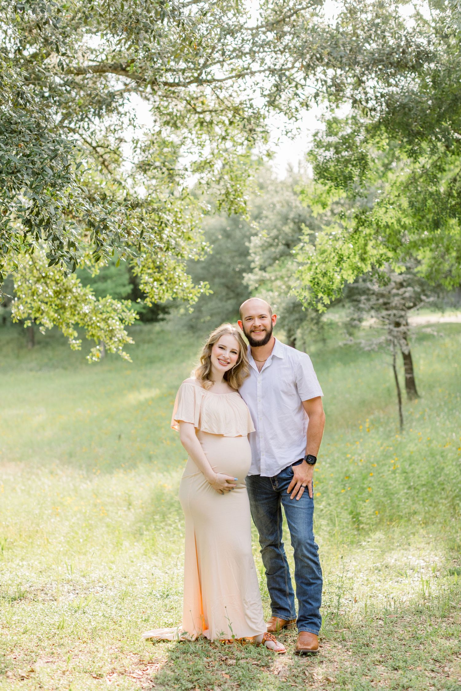 Natural maternity portrait in a sun-dappled meadow with green trees.
