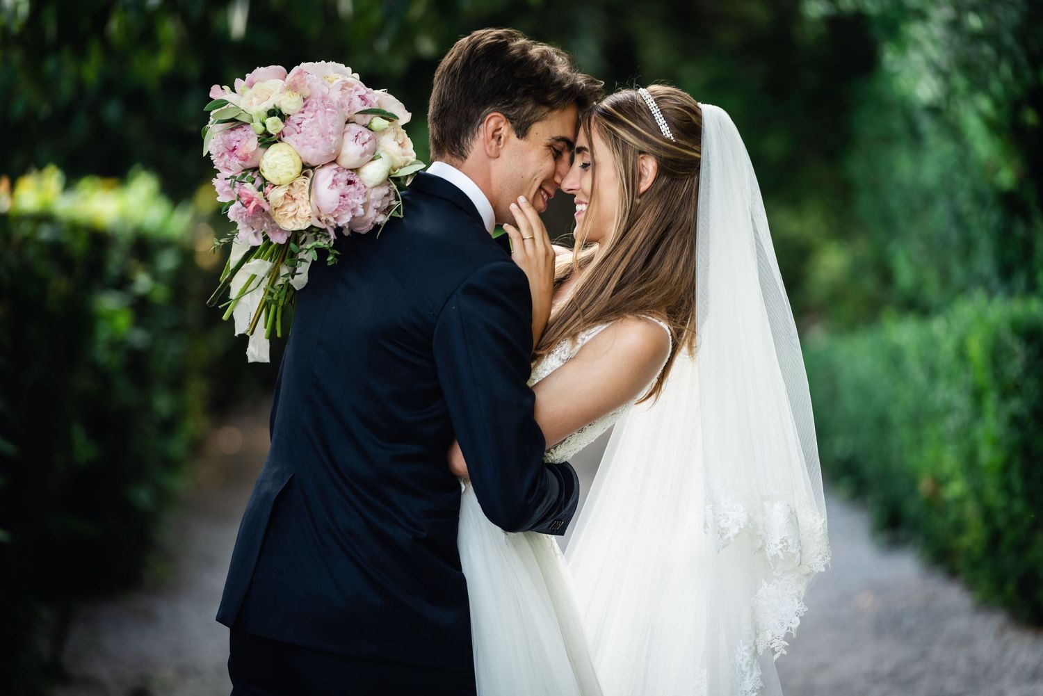 Romantic wedding couple embraces on garden path surrounded by greenery.