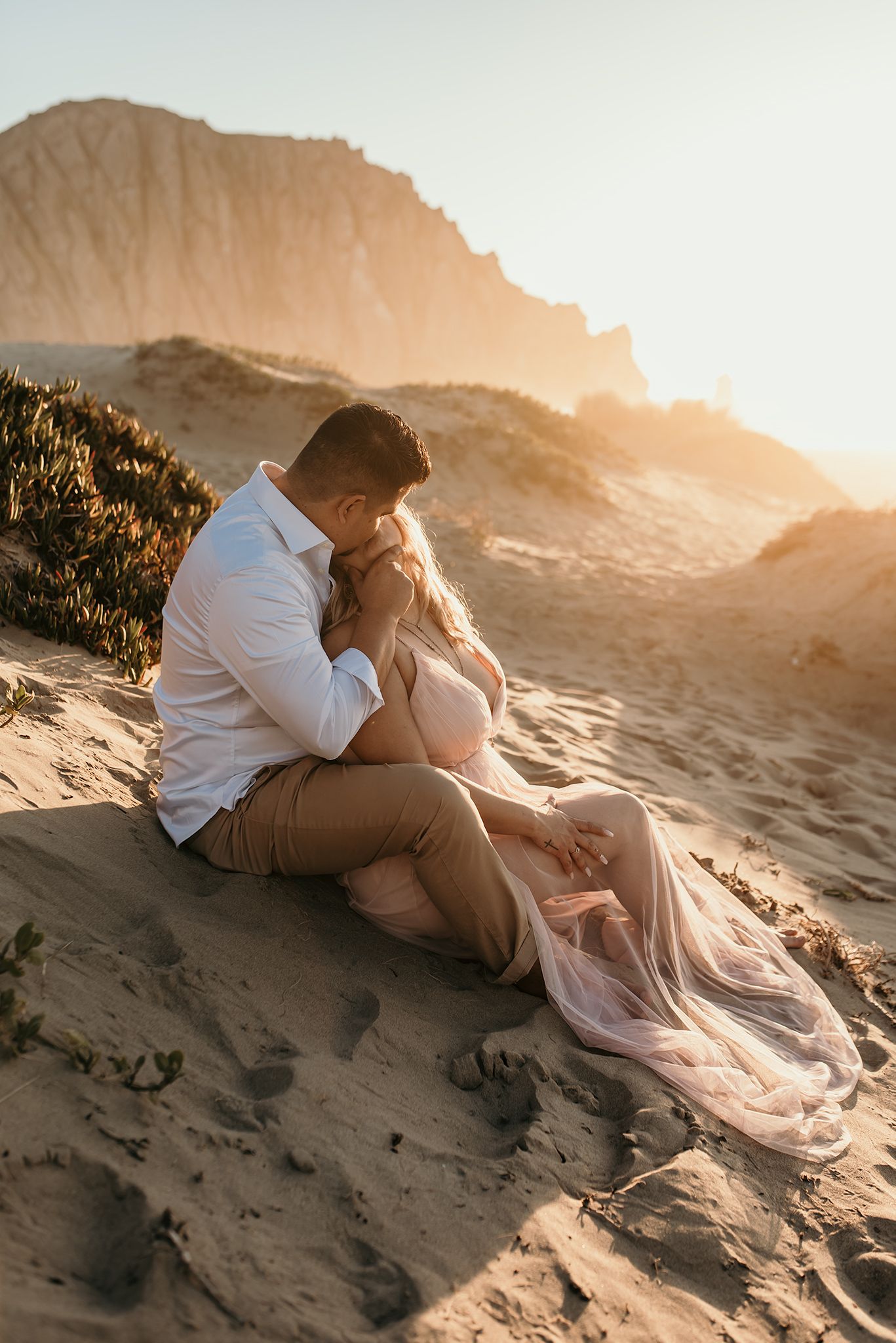 A romantic couple embraces on a sandy hillside during sunset with a dramatic mountain backdrop.