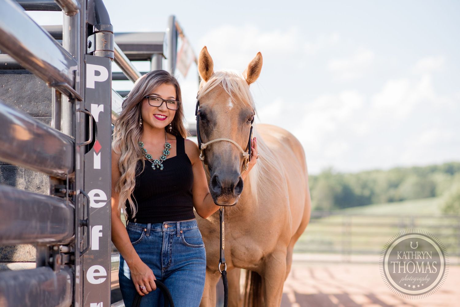 A palomino horse and someone in casual western wear pose together near metal horse stalls.