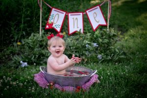 Baby splashes in small tub outdoors with red banner decoration in background.