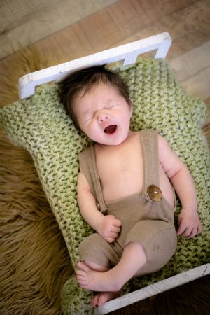 Baby in tan romper yawns while lying on green knit blanket.