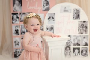 Joyful baby in pink dress smiling while standing next to a decorative column with photo collage background.