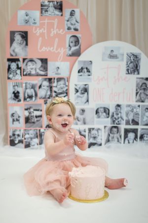 Happy baby in tulle dress celebrating with cake surrounded by black and white photos on pink backdrop.