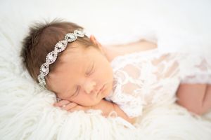 Sleeping newborn baby wearing delicate lace and crystal headband on white fur blanket.