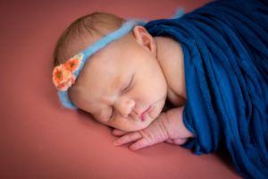Sleeping newborn wrapped in blue fabric with a floral headband against pink background.