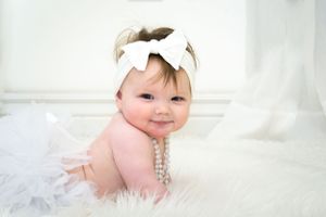 Baby in white bow headband and pearls on furry white rug.