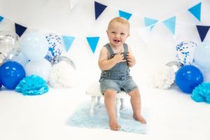 Baby in denim overalls sits with blue and white party decorations and balloons.