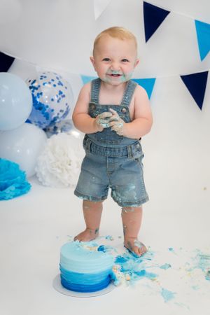 Baby in denim overalls stands on blue cake with party decorations in background.