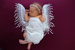 A sleeping newborn lying on burgundy fabric wearing delicate white lace angel wings.