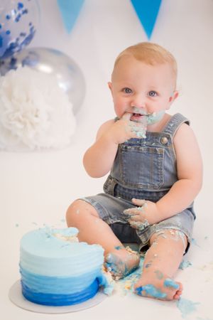 Baby in denim overalls enjoys smashing blue ombre cake during cake smash photoshoot.