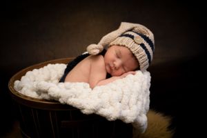 Sleeping baby in striped hat nestled in wooden bowl with white blanket.