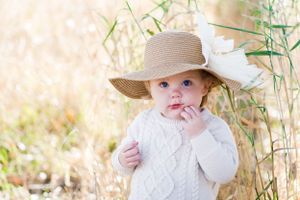 Baby wearing white sweater and straw hat stands among tall grass.