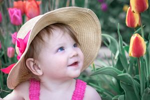 Smiling baby wearing straw hat with pink bow amid blooming orange and pink tulips in garden.