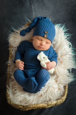 Sleeping newborn in blue knit outfit with matching hat holding small teddy bear on furry backdrop.