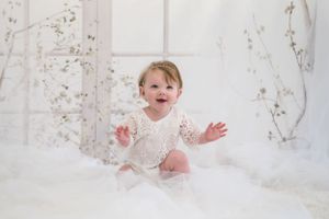 Laughing baby in white lace outfit sitting on fluffy white backdrop with delicate branch decorations.