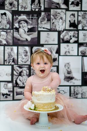 Baby in pink dress excitedly reaches for cake during first birthday photoshoot.