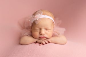 A sleeping newborn baby wearing a white headband rests peacefully on a pink background.