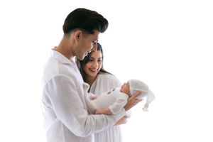 A couple dressed in white holds and gazes lovingly at their newborn baby together against a white backdrop.