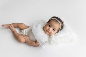 Baby wearing white lacy dress and headband lies on soft white backdrop during professional photoshoot in cedar park tx