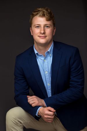 Professional studio portrait against dark background wearing navy blazer, light blue shirt and khaki pants.