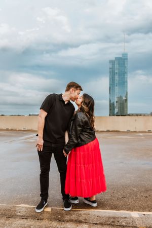 A couple shares a romantic moment on a rooftop with a modern glass skyscraper visible in the cloudy background during a photoshoot in austin