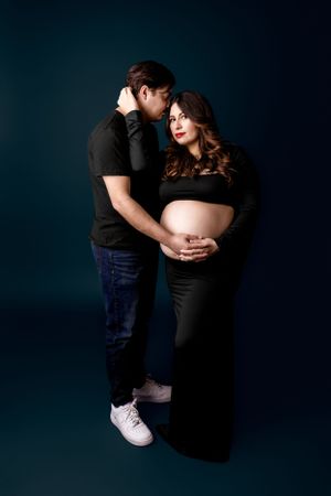 Intimate maternity photo of couple in black clothing against dark background showing tender embrace and bare pregnant belly.